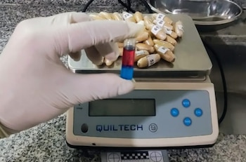 Primer plano de una mano enguantada sujetando una ampolla con líquido rojo y azul, sobre una balanza digital con múltiples cápsulas de color beige etiquetadas
