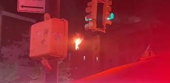 Edificio en llamas de noche, con fuego visible desde una ventana. Semáforos verdes y una caja de control de calle en primer plano, con un resplandor rojo