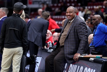 Stacey King, un hombre afroamericano de mediana edad, sonríe, vestido con un traje a cuadros y suéter marrón, sentado al borde de una cancha de baloncesto