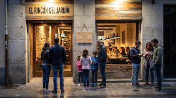 Fachada iluminada del restaurante El Rincón del Jardín, con un cartel 'SOLO CON RESERVA', y diez personas, adultos y niños, esperando afuera.