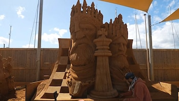 Escultor trabajando en una gran estatua de arena de un rey de ajedrez. Otras piezas de ajedrez de arena y un paisaje de vallas y mástiles de barcos visibles