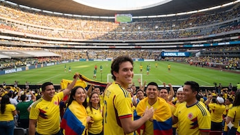 Grupo de personas en un estadio con camisetas amarillas y banderas de Colombia, sonriendo. En el fondo, gradas llenas, jugadores en el campo y pantallas con "Mundial 2026"