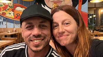 Hombre con gorra negra y mujer de cabello rojizo, ambos sonrientes. Fondo de restaurante con mesas de madera y mural de pizza.