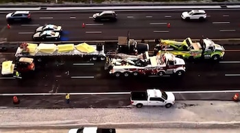 Vista aérea de un accidente en una autopista con dos coches deportivos, un camión de plataforma volcado con carga amarilla, dos grúas y vehículos de asistencia