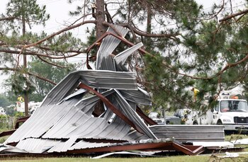 Escombros metálicos retorcidos de una estructura sobre el césped, con un árbol pino dañado en el centro. Al fondo, trabajadores y camiones de servicio