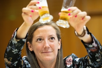 Primer plano de una mujer de cabello castaño que sostiene dos vasos con cerveza rubia por encima de su cabeza. Lleva blusa floral y uñas rosas