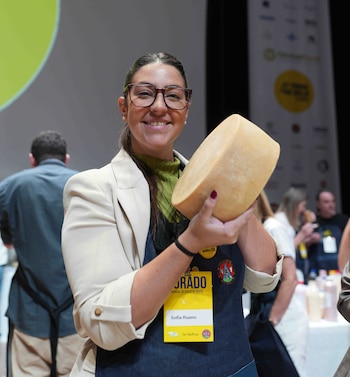 Mujer con gafas, chaqueta beige y delantal azul sonríe mientras sostiene una rueda de queso de color claro con ambas manos. Lleva una credencial de "JURADO"