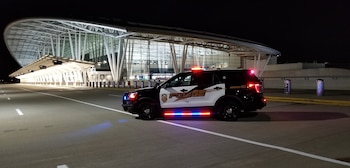 SUV policial negro y blanco con luces rojas y azules intermitentes estacionado de perfil en una carretera oscura frente a la entrada iluminada de un aeropuerto moderno por la noche