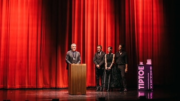 Cuatro personas en un escenario con telón rojo; un hombre habla en un podio de madera y un stand morado con el logo Tip Toe está a la derecha