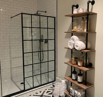 Baño con azulejos blancos, ducha con mampara de cristal negro y estantería industrial de madera y metal llena de toallas y productos de baño, sobre suelo geométrico.
