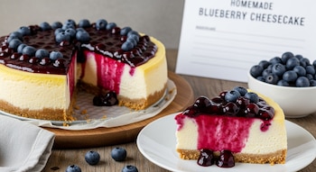 Cheesecake de arándanos cremoso, con una porción cortada en primer plano. Cubierto con salsa de arándanos y bayas frescas. Fondo con más arándanos y cartel.