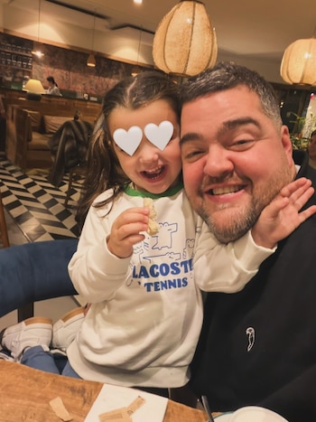 Un hombre y una niña sonríen a cámara en primer plano. Los ojos de la niña están cubiertos por dos corazones blancos. Fondo de restaurante