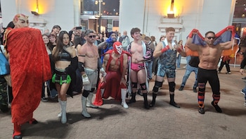 Siete personas, incluyendo un personaje de diablo rojo y seis luchadores con atuendos variados, posan en un área pavimentada frente a un edificio