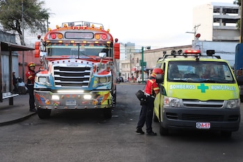 Autobús de pasajeros rojo y azul con decoraciones, una camioneta verde lima de Bomberos Municipales, y dos personas en uniforme de emergencia en una calle