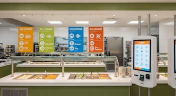 Barra de autoservicio con bandejas de comida, carteles colgantes de alérgenos, quiosco digital táctil y vajilla apilada en restaurante moderno.