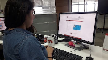 Mujer de perfil frente a monitor de computadora. Sus manos sobre un teclado, un ratón cerca. En la pantalla, página web con formulario e identificación. Taza y lata en el escritorio.