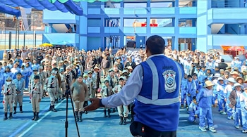 Un hombre de espaldas con un chaleco azul de Sucamec se dirige a decenas de escolares formados en el patio de un colegio