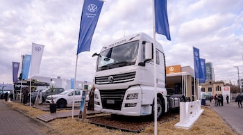Camión blanco Volkswagen Meteor, con logo de la marca. Banderas azules y blancas de Volkswagen. Camioneta pickup Amarok. Personas, stands y cielo nublado