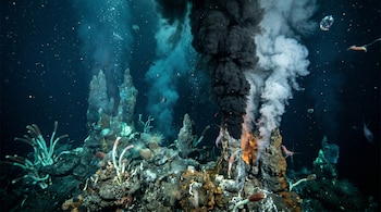 Fotografía submarina de una fumarola hidrotermal en el fondo marino, emitiendo humo mineral blanco y negro, rodeada de oscuridad y vida abisal.