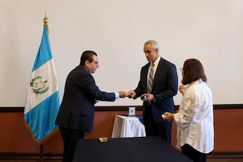 Dos hombres en traje y una mujer junto a una mesa en un salón con la bandera de Guatemala en un costado.