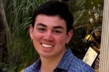 Primer plano de un joven hispano de pelo oscuro sonriendo directamente a la cámara, con un fondo verde borroso de vegetación