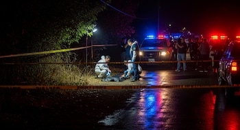 Peritos y policías procesan una escena nocturna con cinta policial amarilla. Se observa un cuerpo en el suelo y vehículos policiales con luces encendidas.