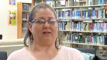 Primer plano de una mujer con gafas moradas y cabello gris atado, usando pendientes largos, frente a estanterías con muchos libros en una biblioteca