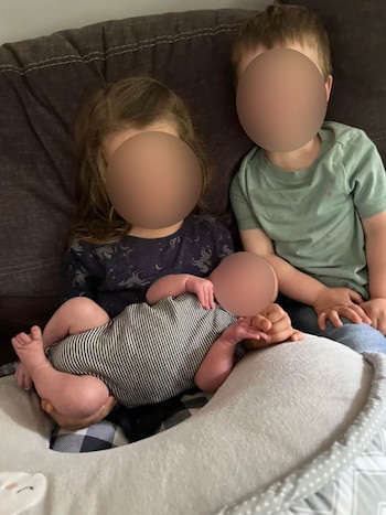 Dos niños y un bebé sentados en un sofá marrón. El bebé, con mameluco a rayas, descansa en un cojín gris. Las caras de los tres están borrosas