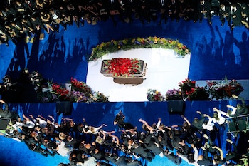 Féretro dorado cubierto de rosas rojas sobre una plataforma blanca, rodeado de arreglos florales, con personas de pie y sentadas en un escenario azul