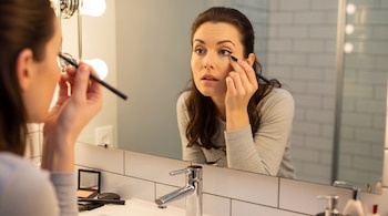 Una mujer maquillándose con delineador frente a un espejo de tocador con luces. Se ven brochas, un compacto de maquillaje y un lavamanos.