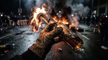 Primer plano de una mano con guante de cuero que sostiene una botella con mecha en llamas, con humo y fuego en el fondo nocturno.