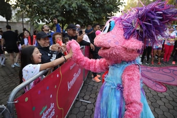 Un personaje peludo de color rosa con vestido azul da la mano a una niña detrás de una valla de protección