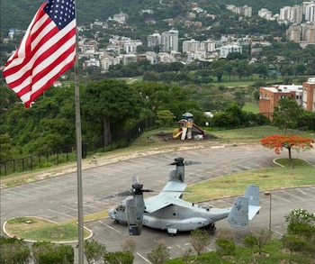 Una bandera de EE. UU. ondea frente a un avión militar V-22 Osprey gris estacionado en un lote; en el fondo, edificios de la ciudad se extienden por colinas verdes