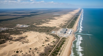 Vista aérea de una larga costa con océano, olas, dunas de arena, llanuras y una gran instalación industrial cerca de la playa bajo un cielo parcialmente nublado.