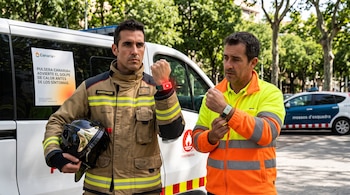 Un bombero con su casco y un trabajador con chaqueta reflectante exhiben las pulseras que llevan en sus muñecas frente a un vehículo oficial.
