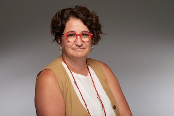 Retrato de Virginia Messi, mujer de piel clara con pelo corto castaño y gafas rojas, vistiendo un chaleco marrón sobre una blusa blanca y un collar rojo