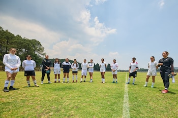 Onze mulheres, vestindo uniformes e chuteiras de futebol, estão em campo. Há árvores e um céu nublado ao fundo