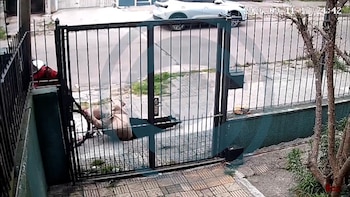 Una reja metálica de una vivienda frente a una persona caída en la acera y un individuo con casco en el exterior