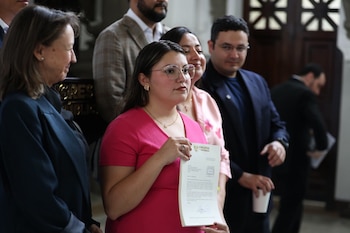 Una mujer con anteojos y blusa rosada sostiene un documento impreso frente a un grupo de personas