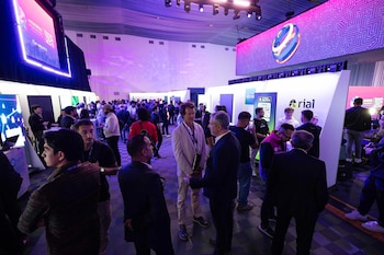 Vista general de un concurrido evento de negocios con personas interactuando en un espacio iluminado con luces moradas, pantallas LED y stands de empresas