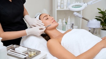 Mujer con toalla y diadema en camilla. Especialista con guantes realiza tratamiento facial con bisturí. Bandeja con utensilios y productos. Lámpara.