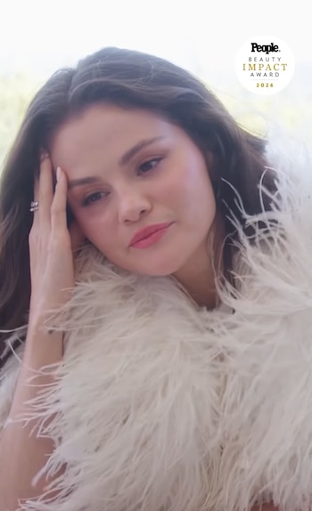 Selena Gomez apoya la mano derecha en la sien mientras viste una prenda cubierta de plumas blancas