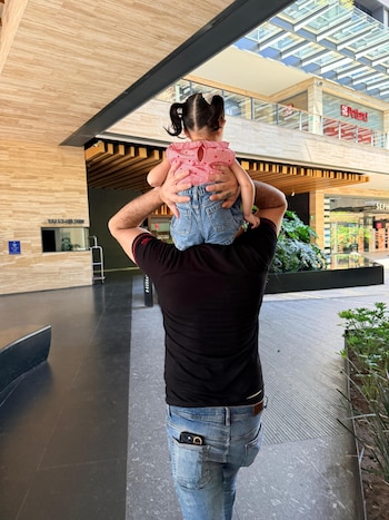 Un hombre de espaldas lleva a una niña pequeña en sus hombros dentro de un centro comercial. La niña usa blusa a cuadros rojos y jeans