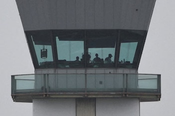 Vista exterior de la parte superior de una torre de control de tráfico aéreo con grandes ventanas. Tres figuras de personas se ven silueteadas dentro de la cabina de control