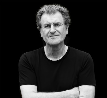Retrato en blanco y negro de Alfredo Jaar, un hombre con cabello rizado gris, gafas, camiseta negra, y los brazos cruzados, mirando directamente a la cámara sobre fondo oscuro