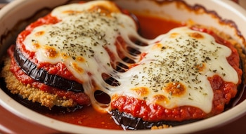 Primer plano de berenjenas apanadas con capas de salsa de tomate, queso mozzarella fundido y orégano espolvoreado en una fuente de cerámica.