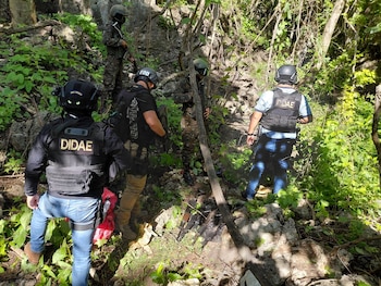 Cinco personas con cascos y chalecos tácticos, algunas con uniforme militar, en un terreno rocoso con vegetación. Varias armas largas en el suelo