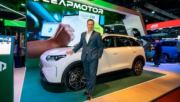 Un hombre con traje posa junto a un automóvil blanco en un escenario con una pantalla luminosa de fondo