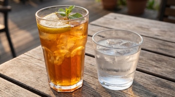 Dos vasos, uno con té helado, rodajas de limón y hojas de menta, y el otro con agua y cubos de hielo, sobre una mesa de madera.