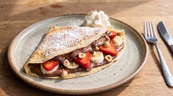 Crepe con chocolate, fresas, banana, avellanas, azúcar glas y crema batida en un plato. Cubiertos al lado sobre mesa de madera.